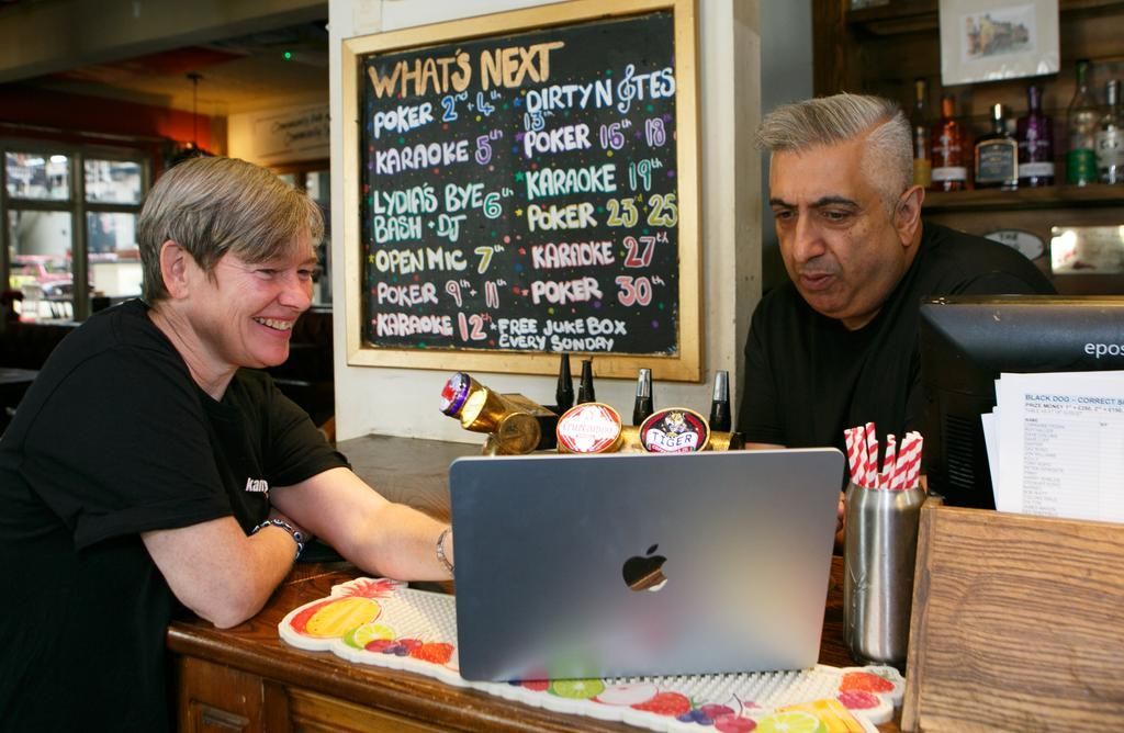 Two people at a bar looking at a laptop. A chalkboard lists upcoming events.