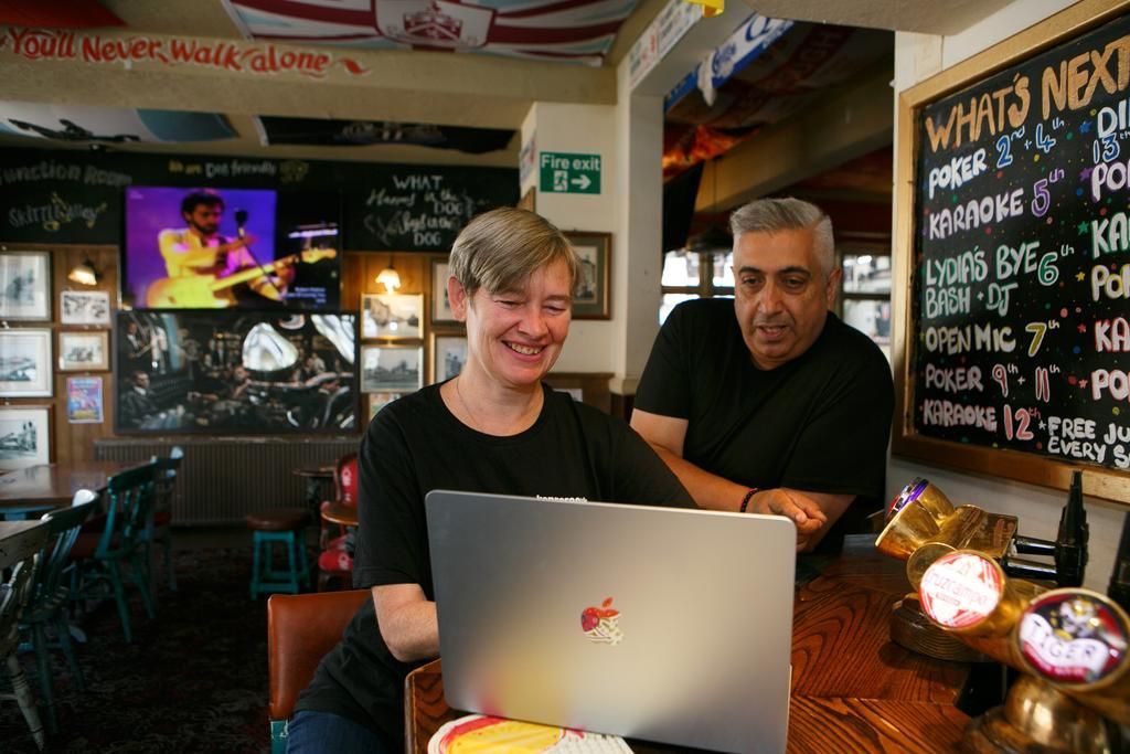 Two people smiling behind a laptop in a cozy café, with chalkboard menus and neon signs in the background