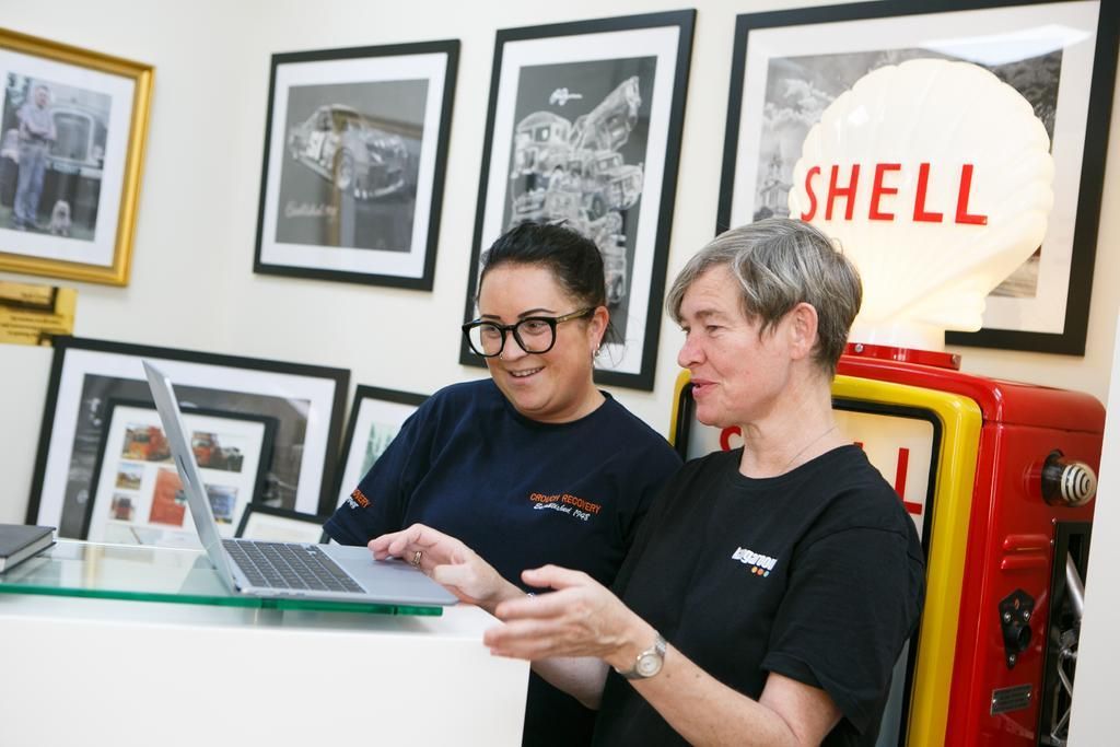 Two women looking at a laptop; in an office with framed art and a Shell gas pump replica.