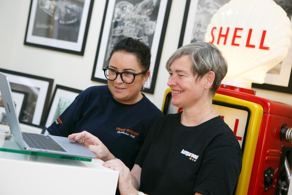 Two women looking at a laptop. They are smiling. A vintage Shell gas pump sign is visible.
