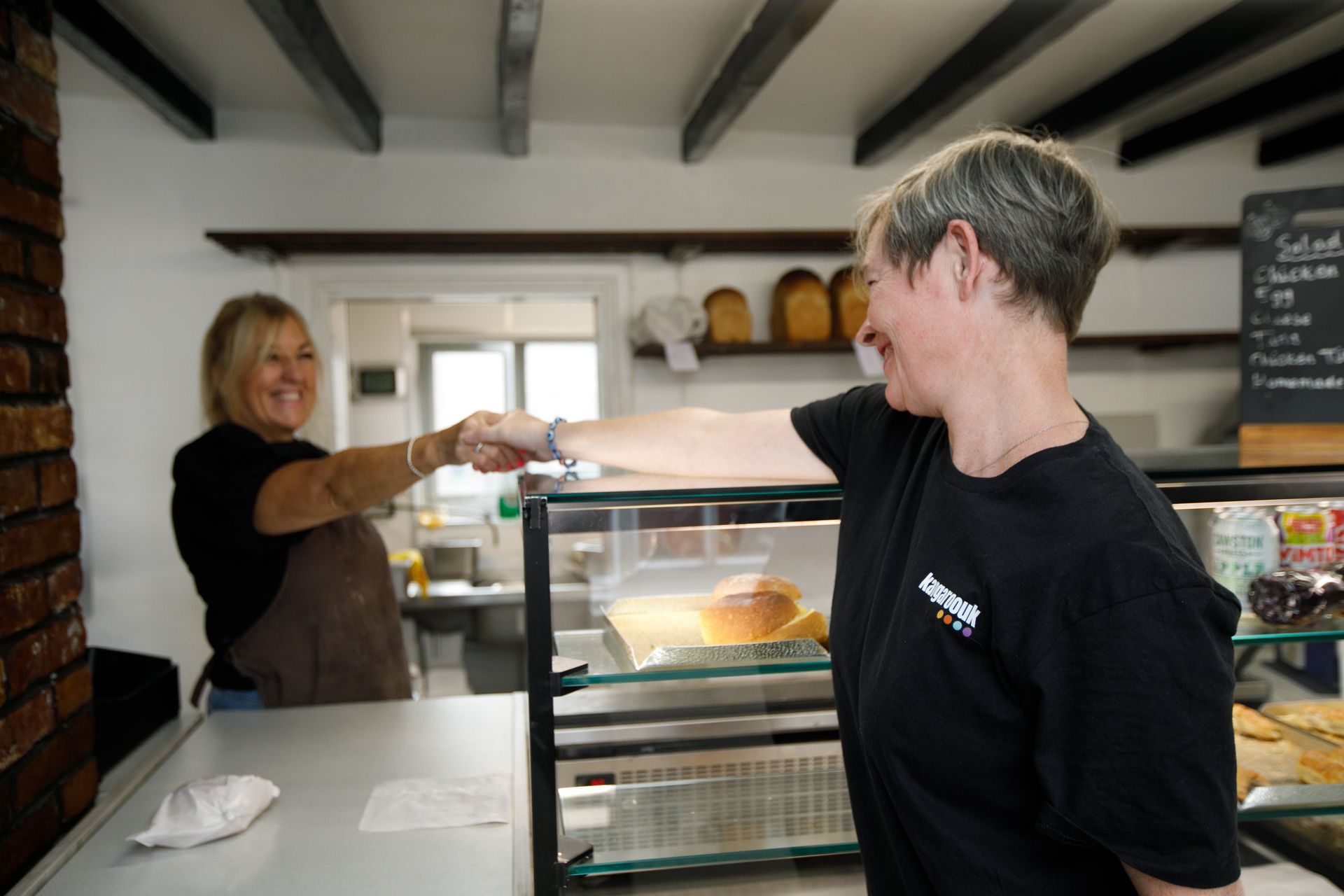 Two women bump fists inside a cafe. One smiles behind the counter, the other smiles while facing the camera.