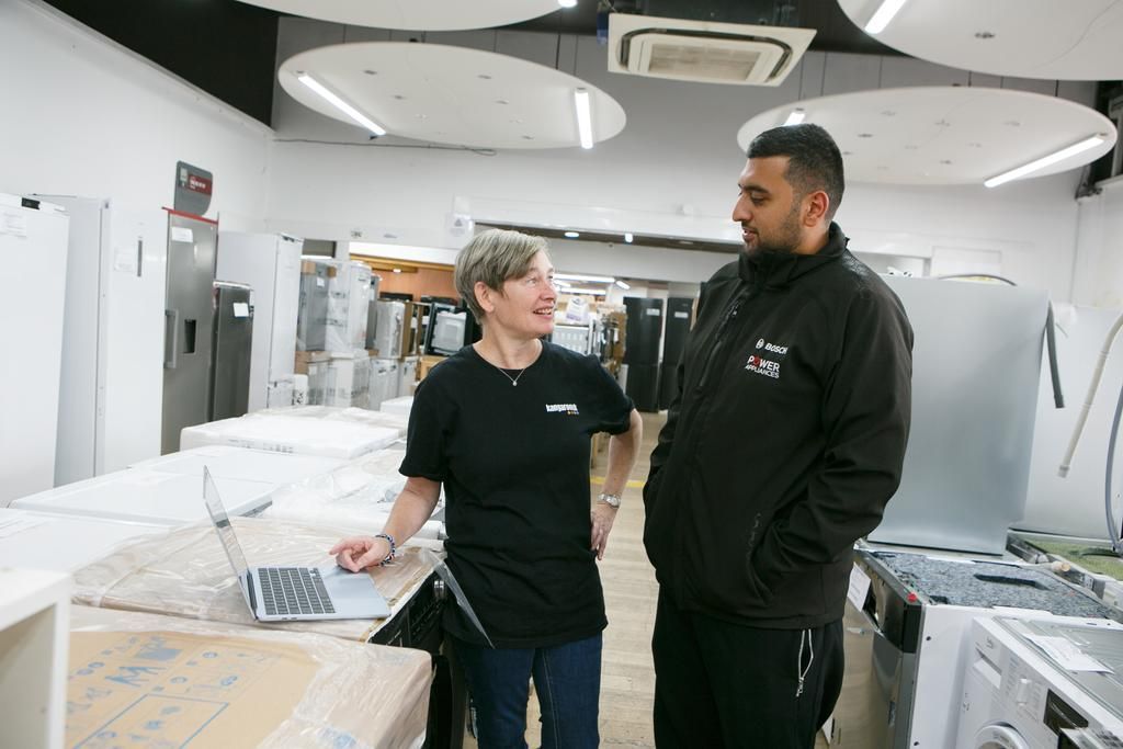 Two people standing in a warehouse, talking while one looks at an open laptop on a workbench near various appliances.