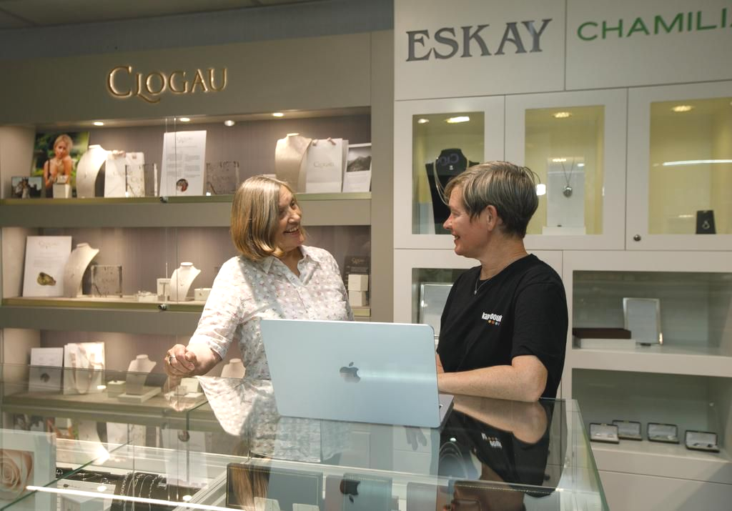 Two women in a jewelry store, smiling at each other. One works on a laptop, the other gestures near a display case.