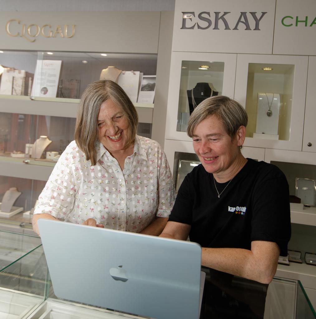 Two women smiling, looking at a laptop in a jewelry store. Shelves of jewelry visible behind them.