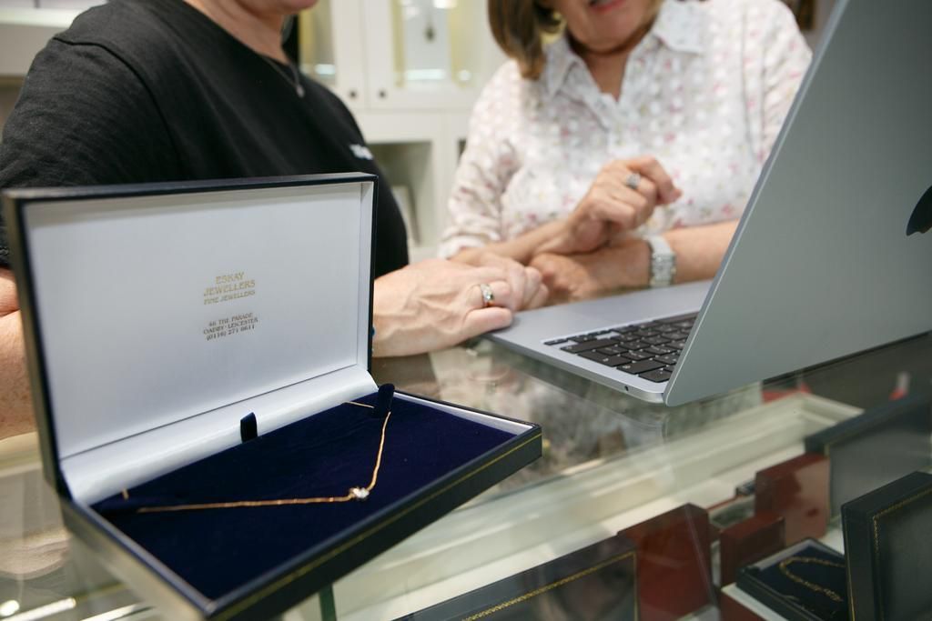Two women examining a diamond necklace in an open box, near a laptop on a jewelry store counter.