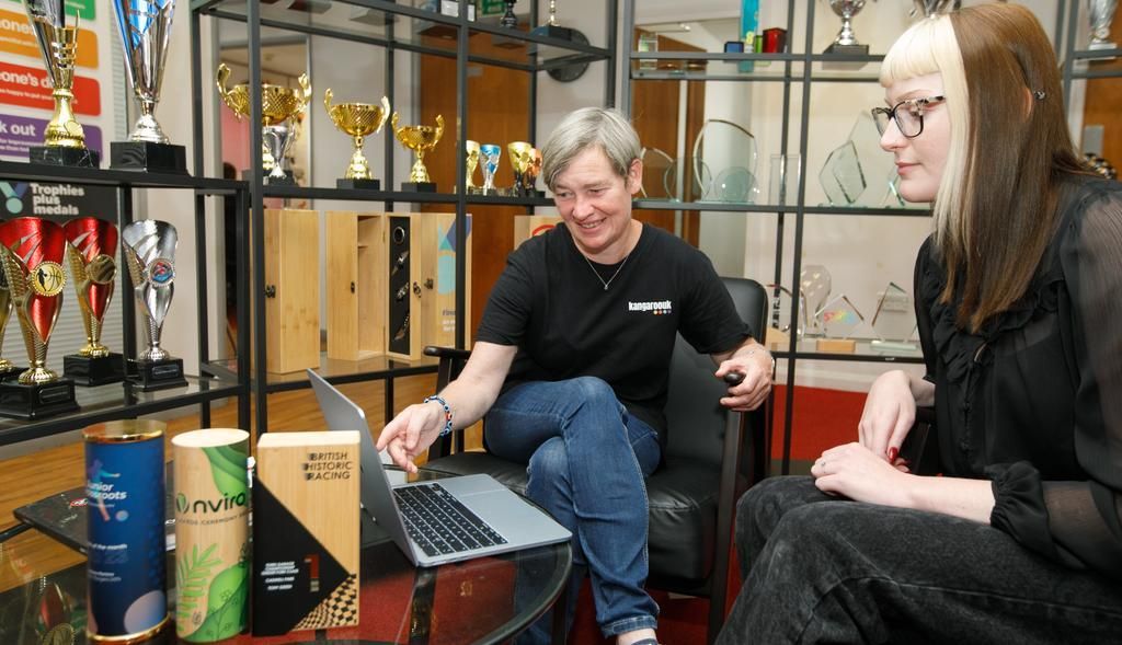 Two women sit by a laptop, looking at the screen. Trophies are on shelves behind them. Tea boxes are on a table.