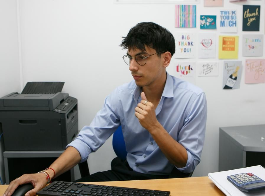 A person in a light blue shirt works at a desk, touching their chin while looking at a computer monitor.