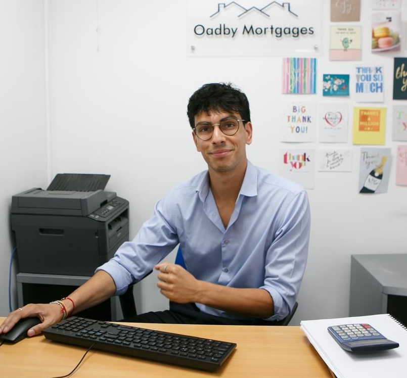 Man at desk in an office. He wears glasses and a blue shirt, smiling at the camera.
