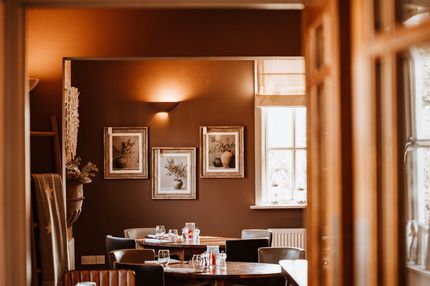 Cozy restaurant dining room with warm brown walls, framed art, and set tables by a window.