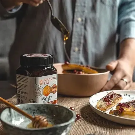 Manuka honey being drizzled over food on a table; jar of honey, wooden honey dipper and bowl are present.