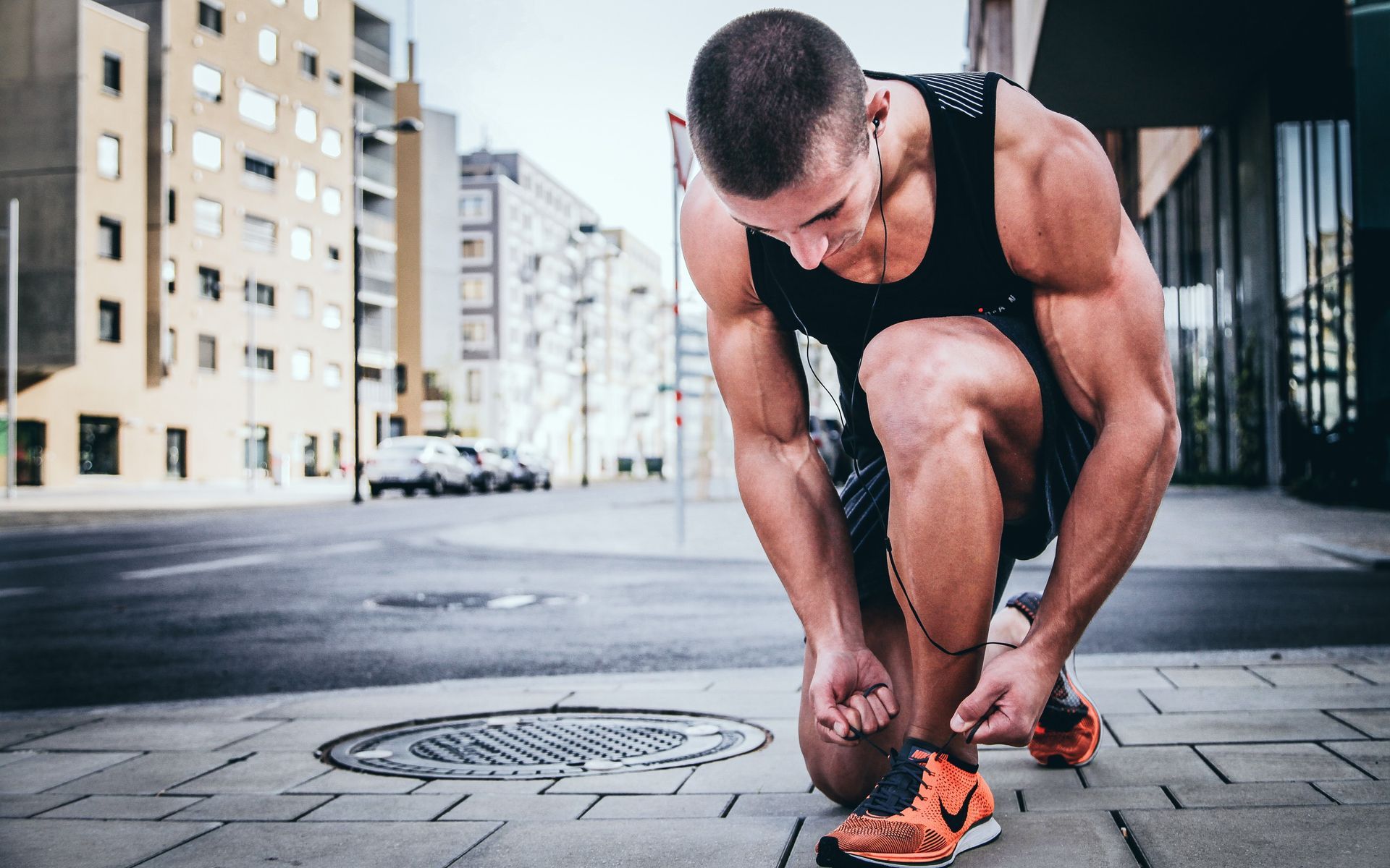 A muscular athlete in a black tank top kneels on a city sidewalk, tying the laces of bright orange sneakers.
