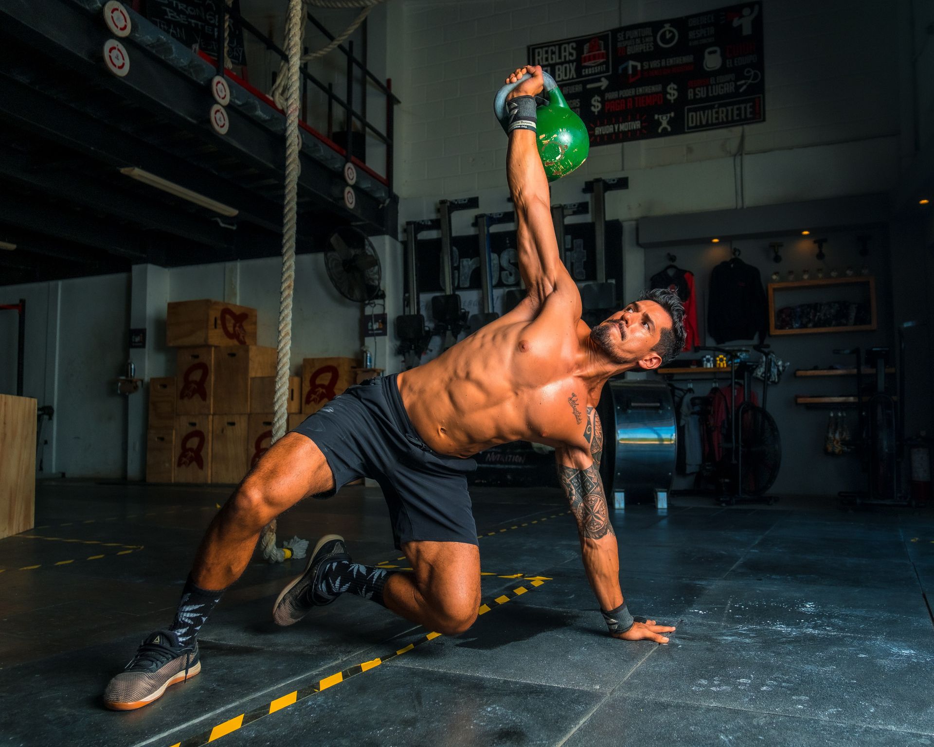 Shirtless athlete performing a kettlebell windmill exercise in a gym with a green kettlebell held overhead.