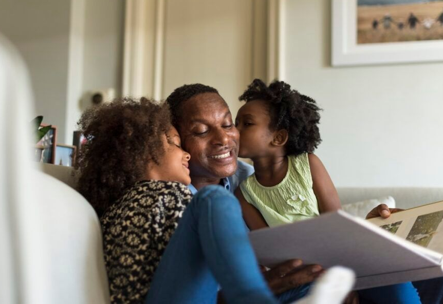 Father and two daughters smiling, looking at a photo album together on a sofa.
