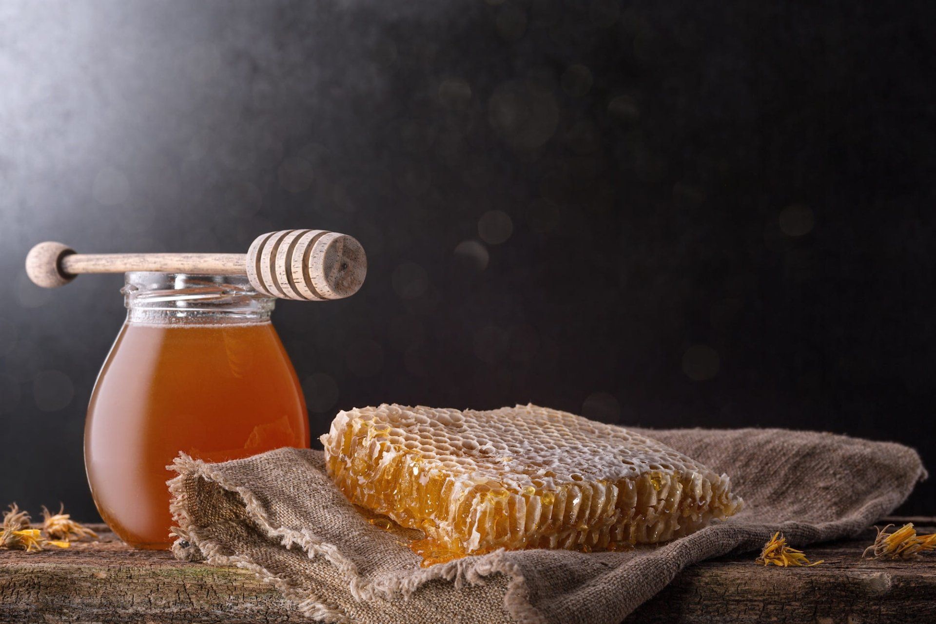 Jar of honey with dipper, honeycomb on cloth. Wooden surface, dark background.