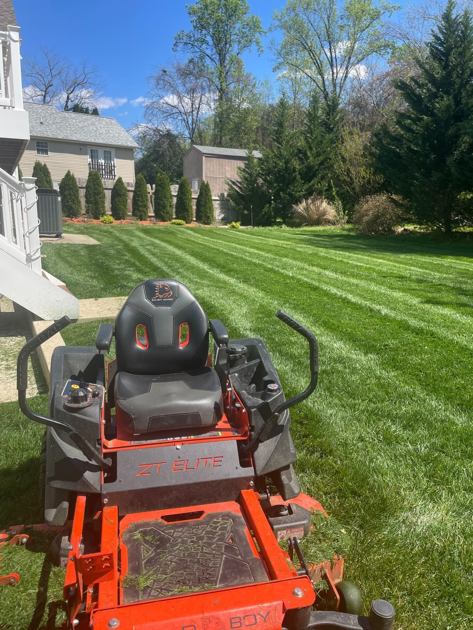 Red riding lawn mower on a striped green lawn beside a house and tall shrubs under a blue sky