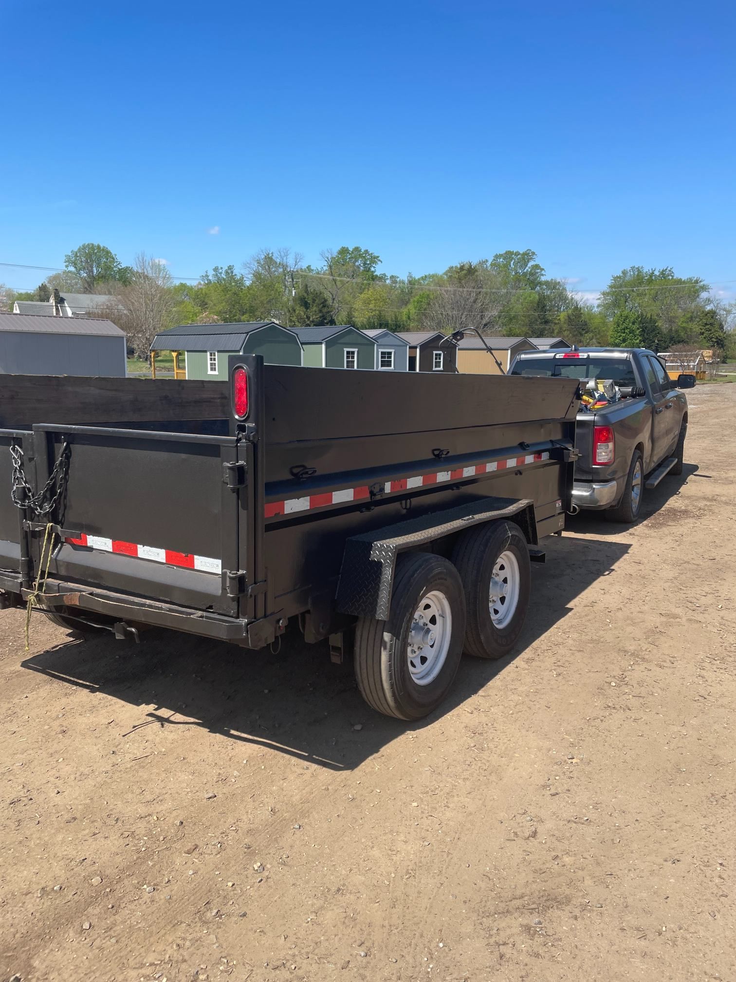 Black dump trailer hitched to a pickup truck parked on a dirt lot under a blue sky.