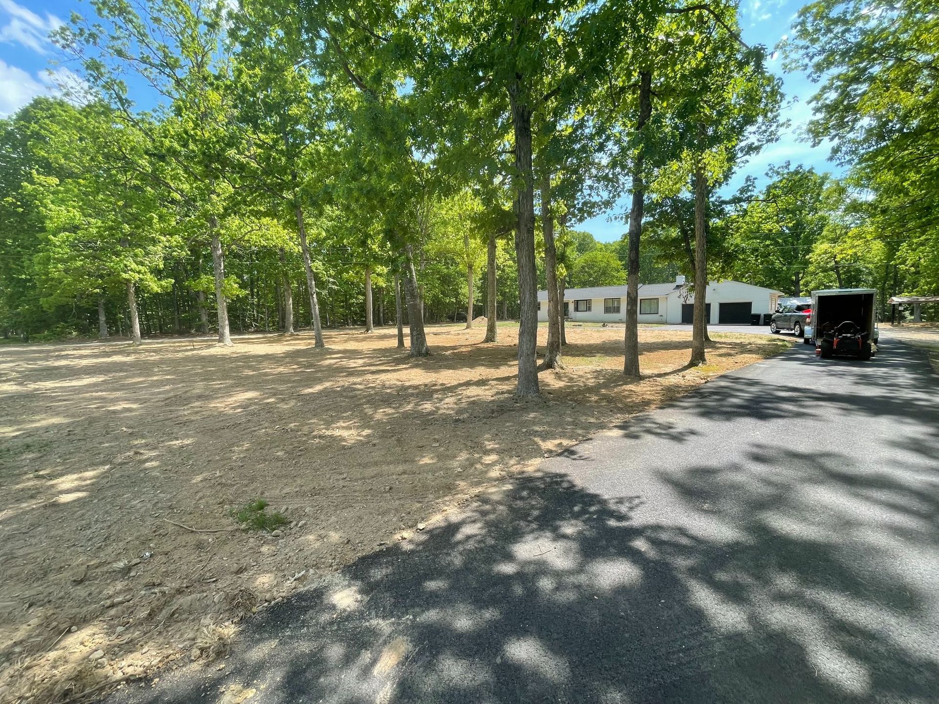 Tree-lined gravel lot beside a road, with a white building and trailer in the background under a sunny sky.