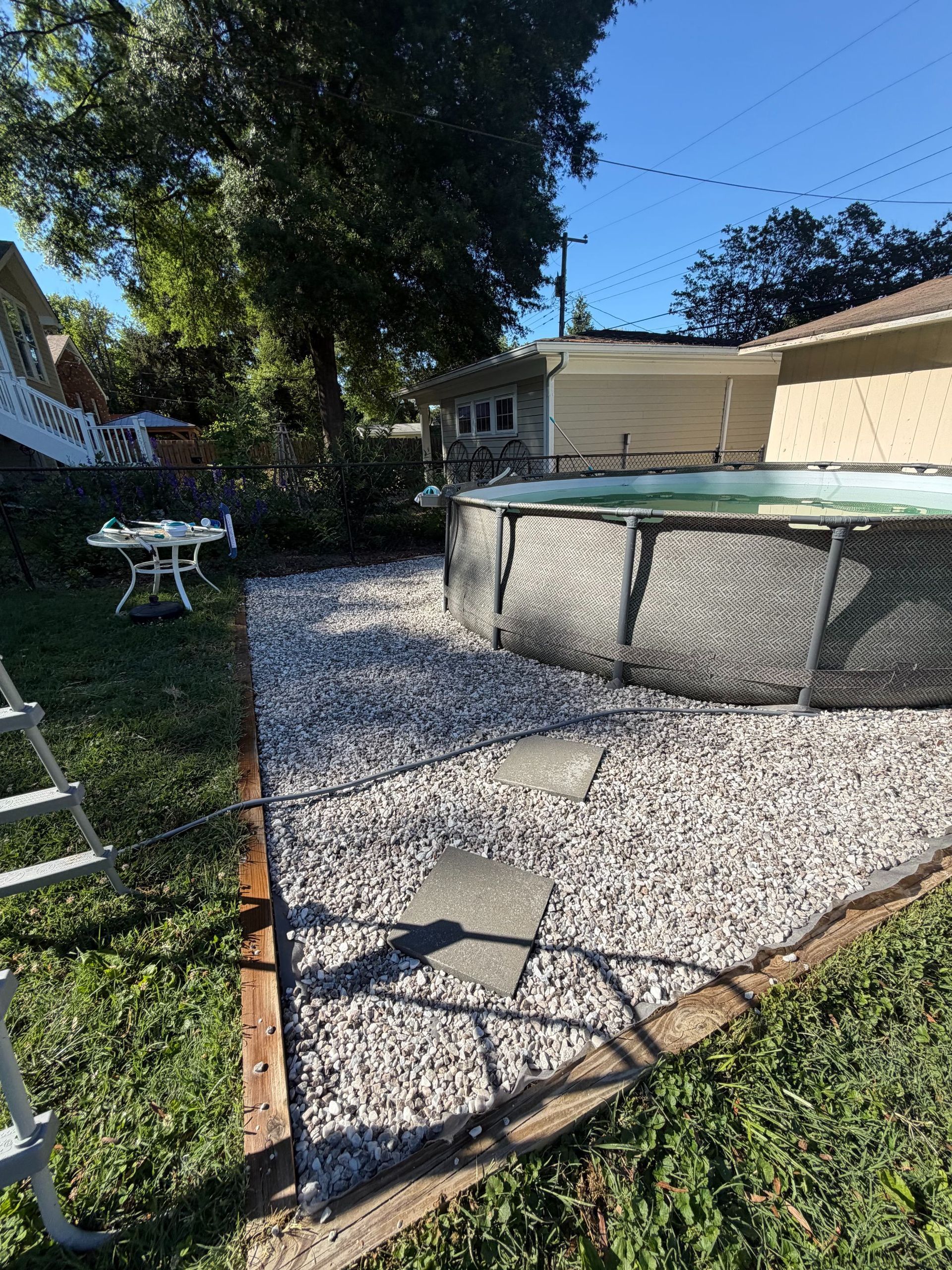 Backyard with gravel area, above-ground pool, picnic table, and ladder beside trees and grass