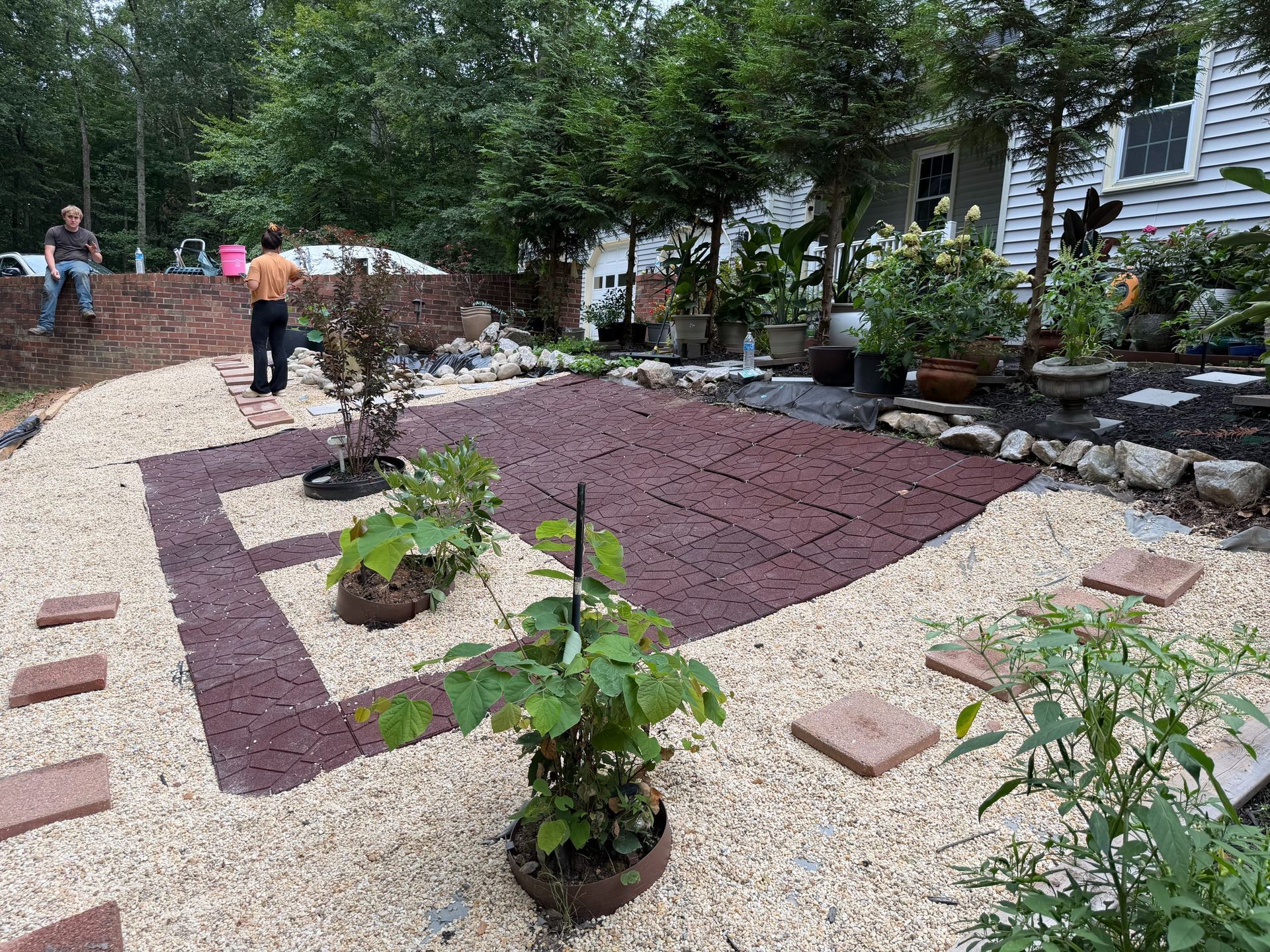 Backyard garden with gravel path, raised red brick bed, and potted plants along a house and trees