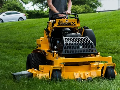 Yellow riding lawn mower cutting grass on a green lawn, with a person standing behind it.