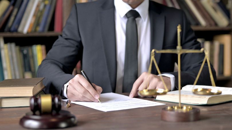 Abogado escribiendo en su escritorio con mazo, libros y balanza de la justicia.