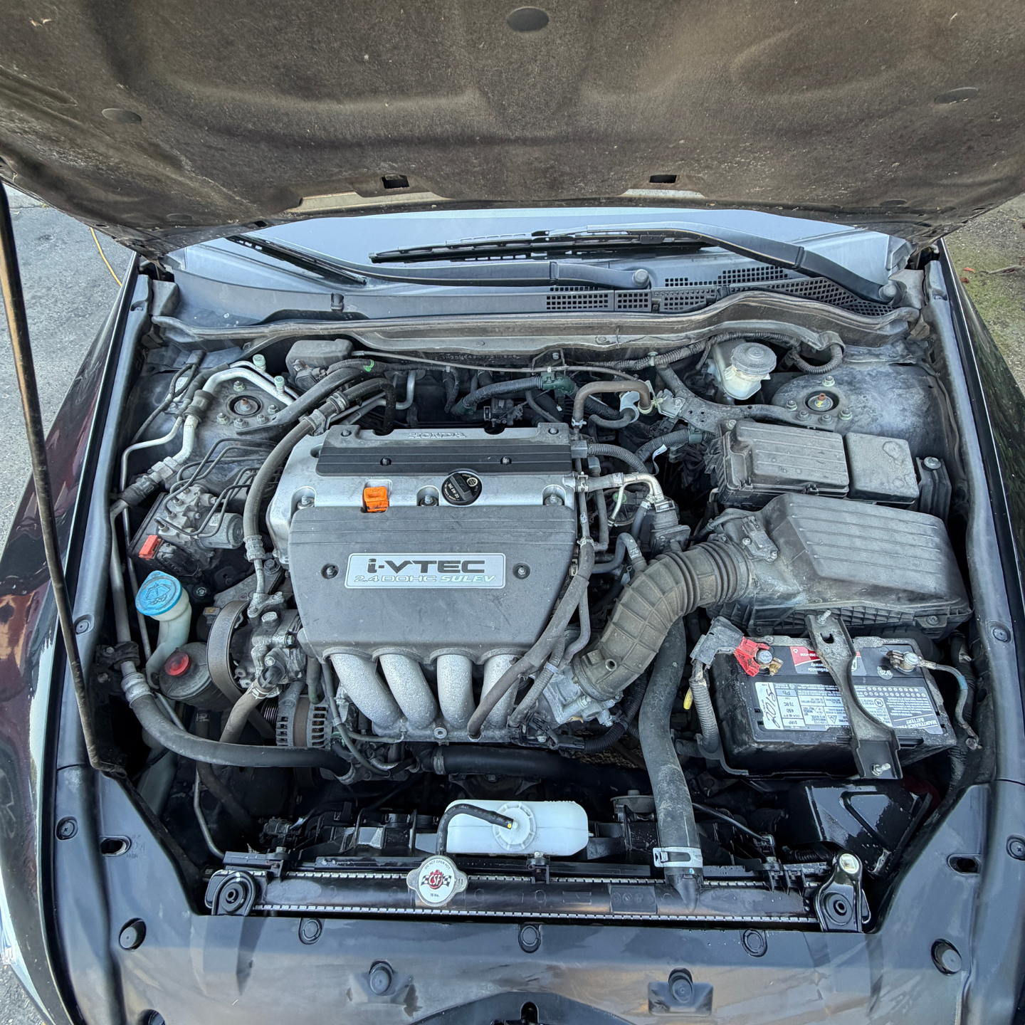 Car engine bay with hood open, showing a silver engine cover and battery on the right.