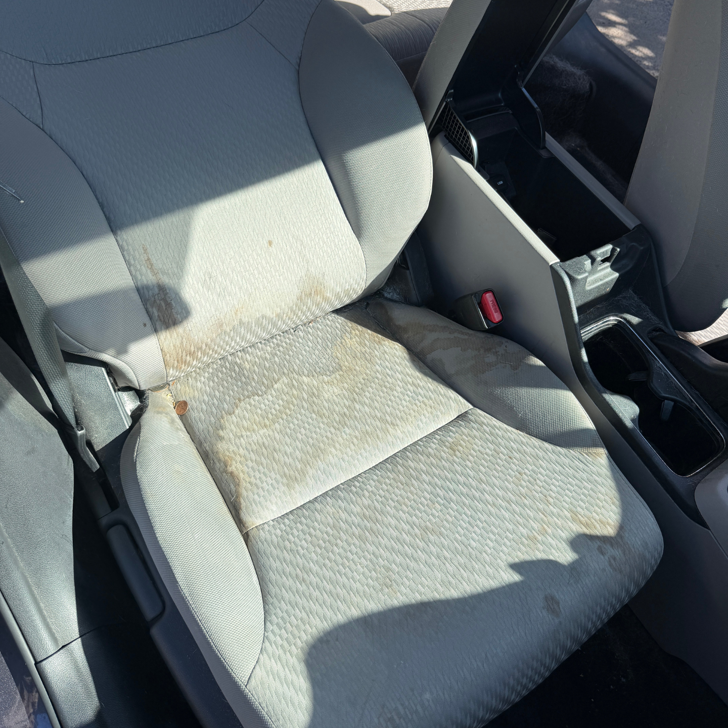 Light gray car front seat with a large stain on the cushion and backrest.