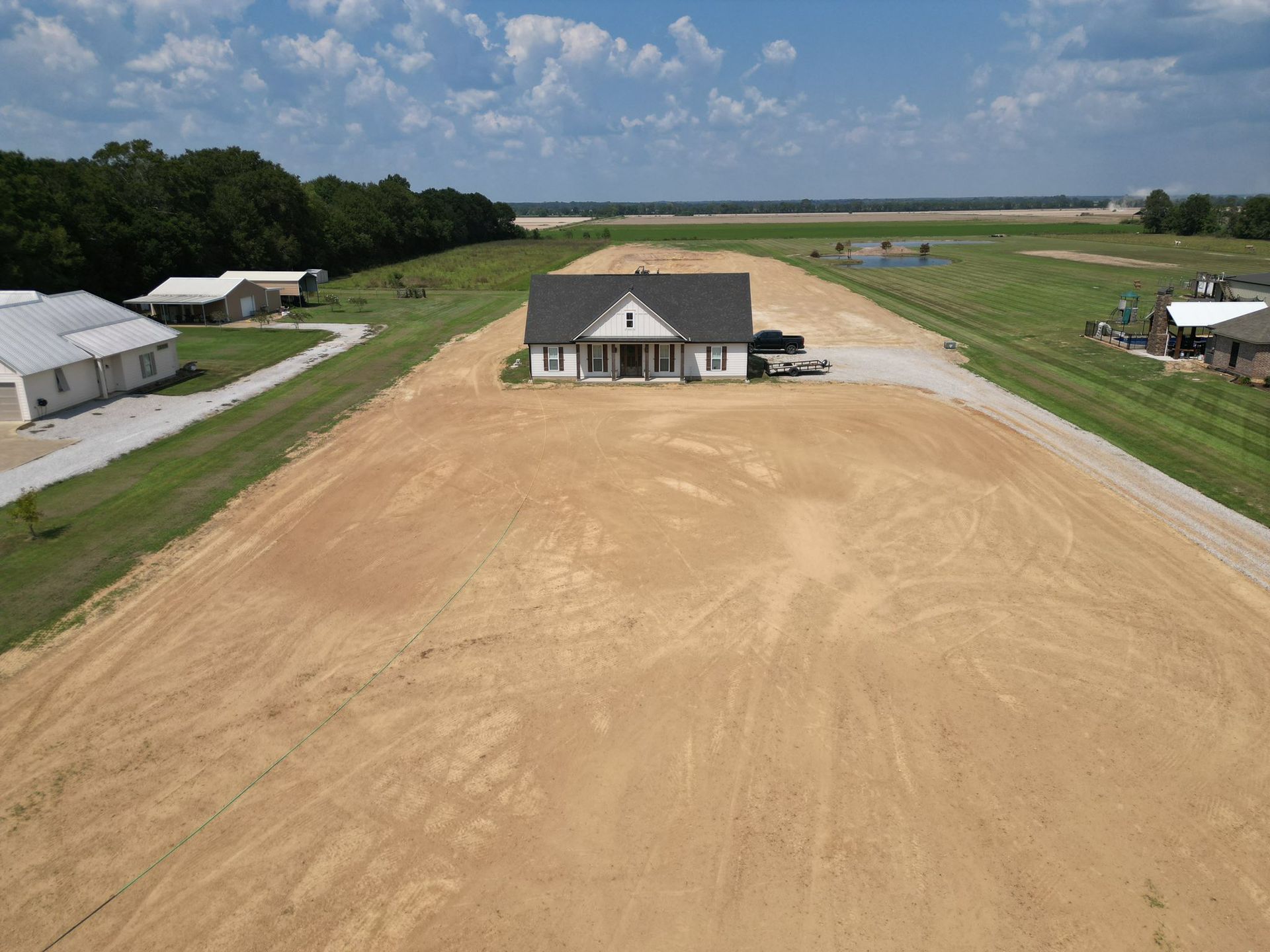 A house under construction on a large cleared lot with neighboring homes and a blue sky.