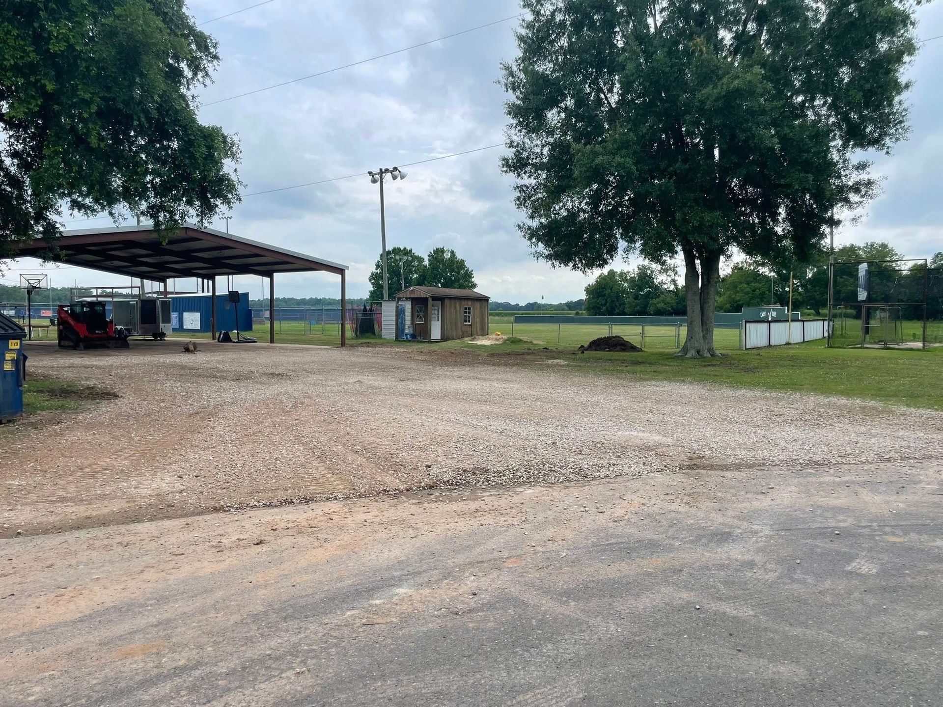 Gravel parking area beside a covered shelter, trees, and open fields under a cloudy sky.
