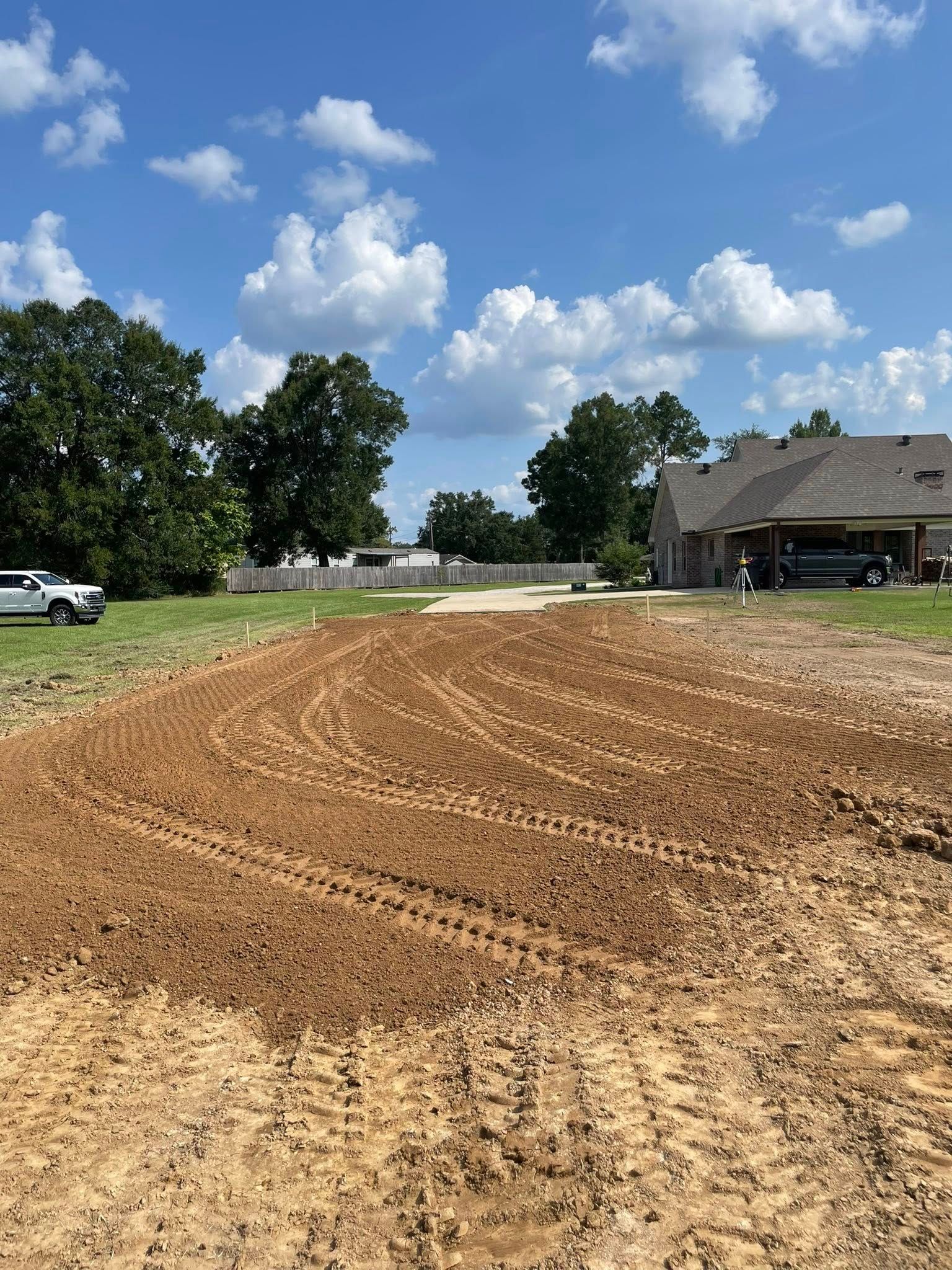 Freshly graded dirt lot beside a house under a bright blue sky with trees and parked vehicles