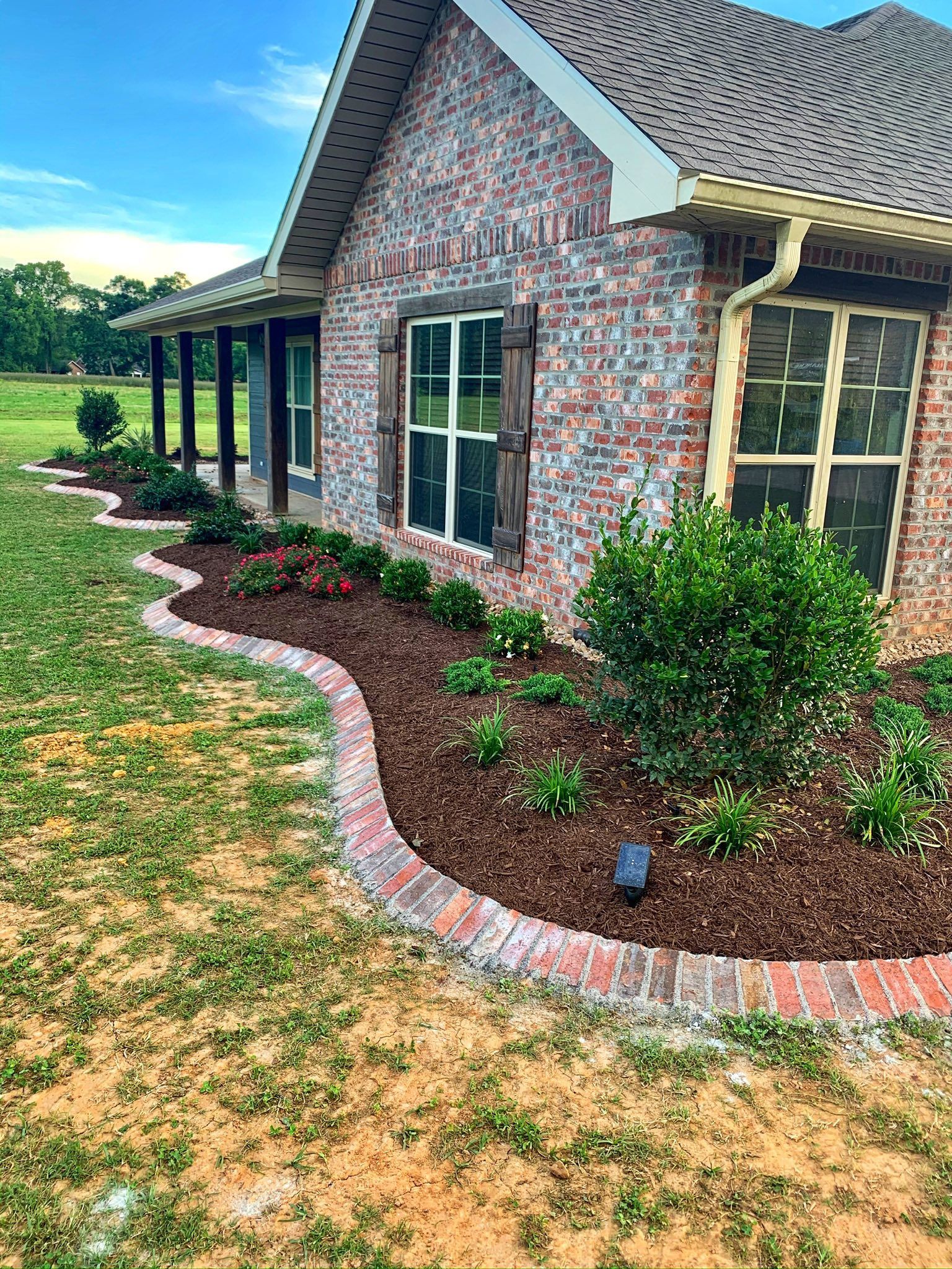 Brick house with dark windows, curved stone-edged mulch bed, and green lawn under a cloudy sky