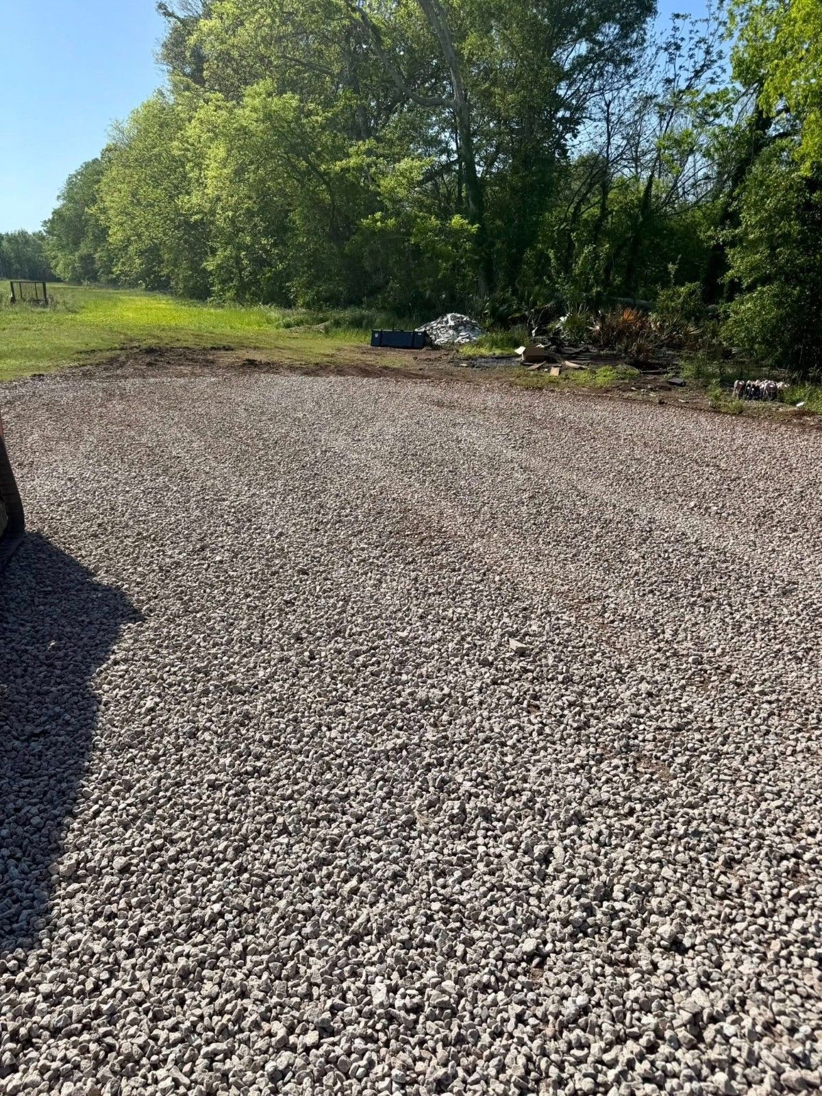 Gravel driveway beside green trees under bright sunlight, with a few parked cars in the distance