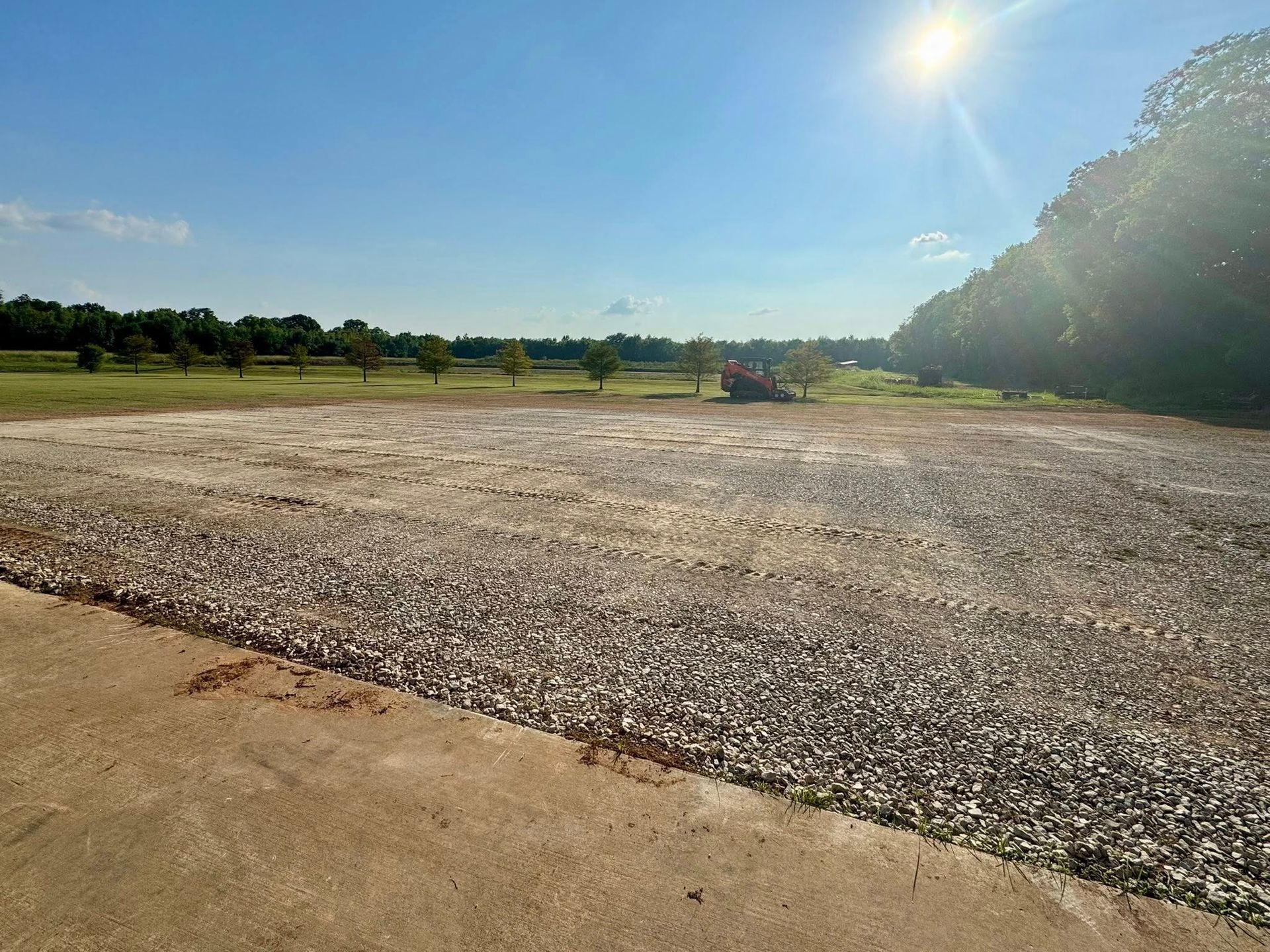 Sunny gravel lot beside grassy field and trees under a clear blue sky