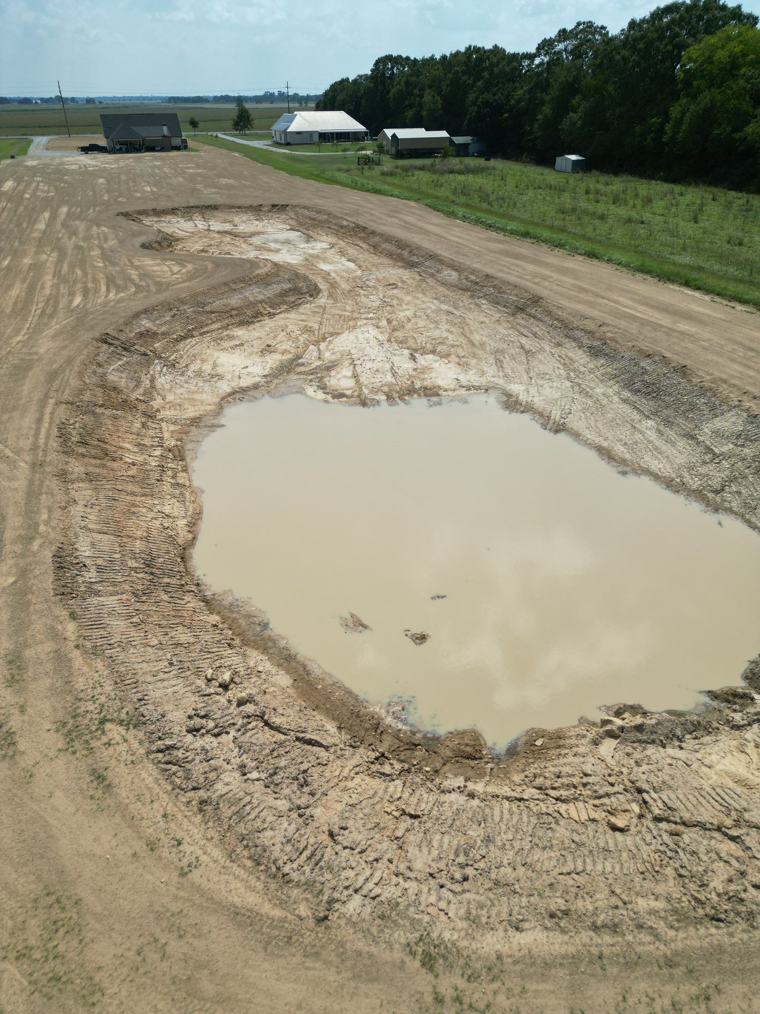 Mud-filled dirt road with a large puddle beside green fields and distant farm buildings