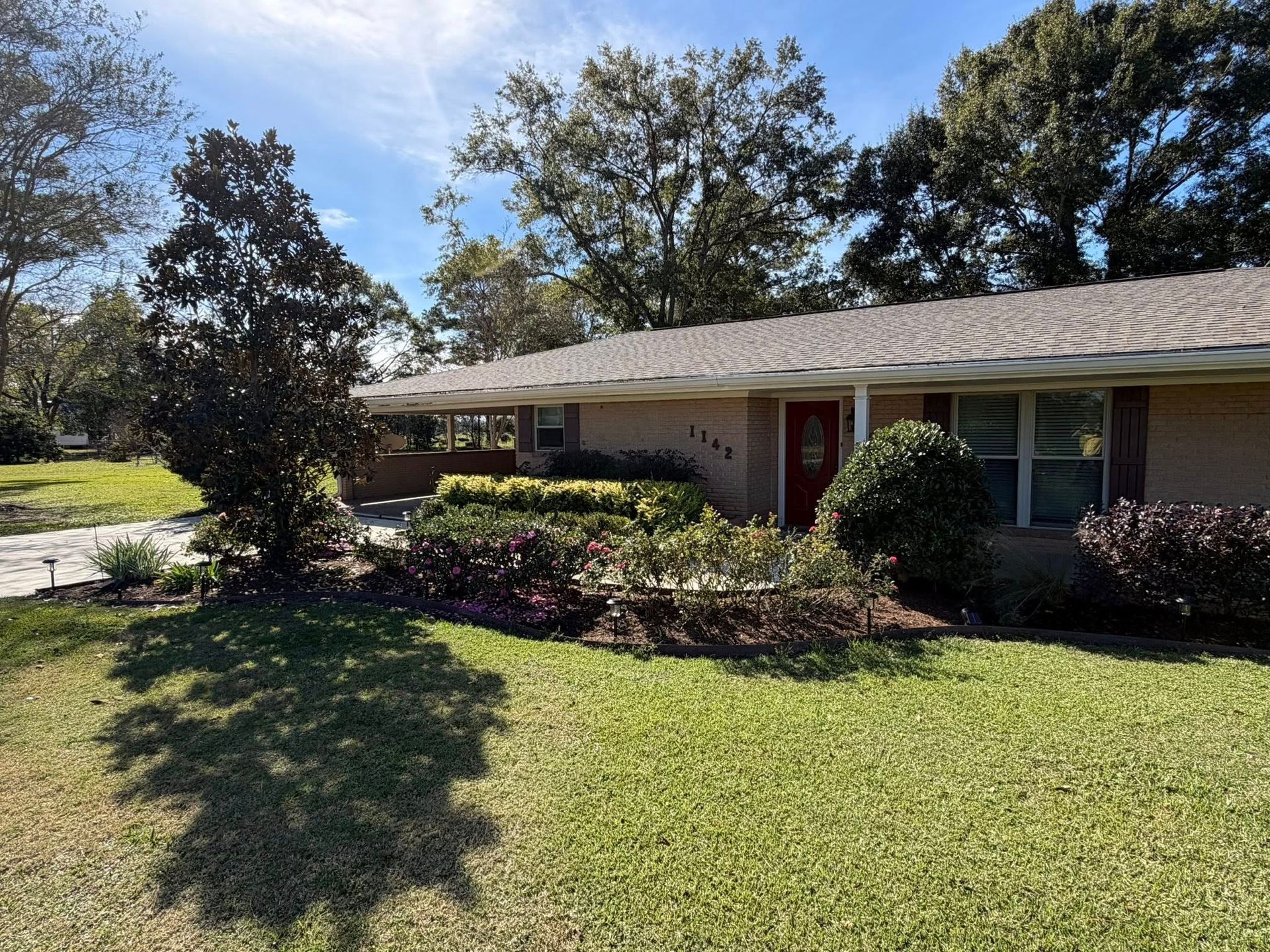 Single-story suburban house with a manicured lawn, trees, and shrubs in front