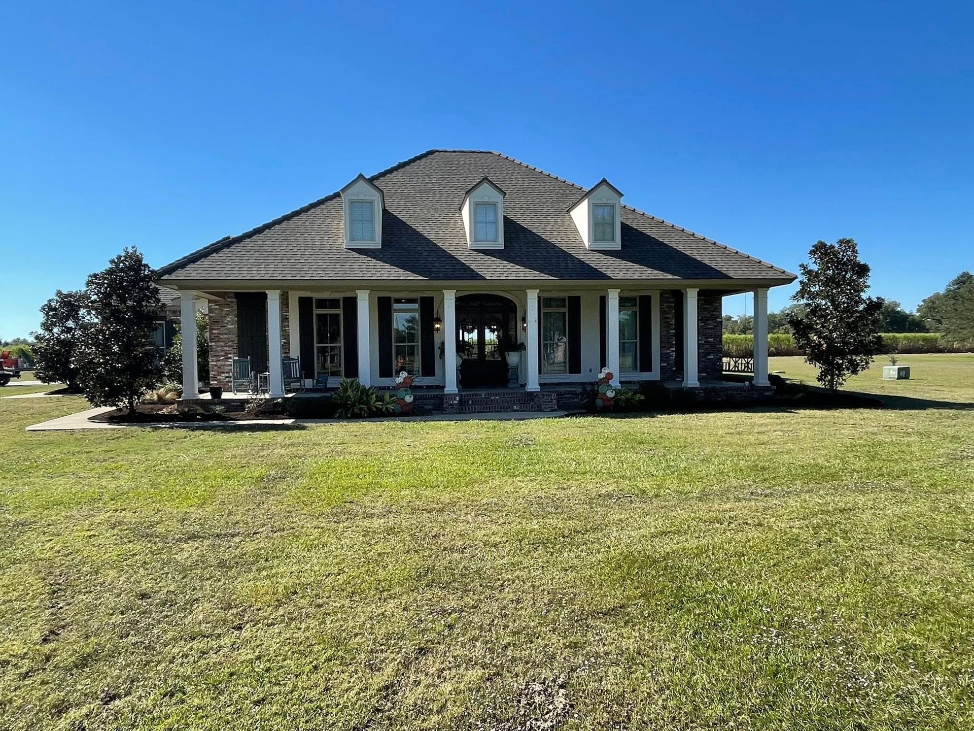 Single-story house with a gray roof and white columns on a large grassy lot under a clear blue sky