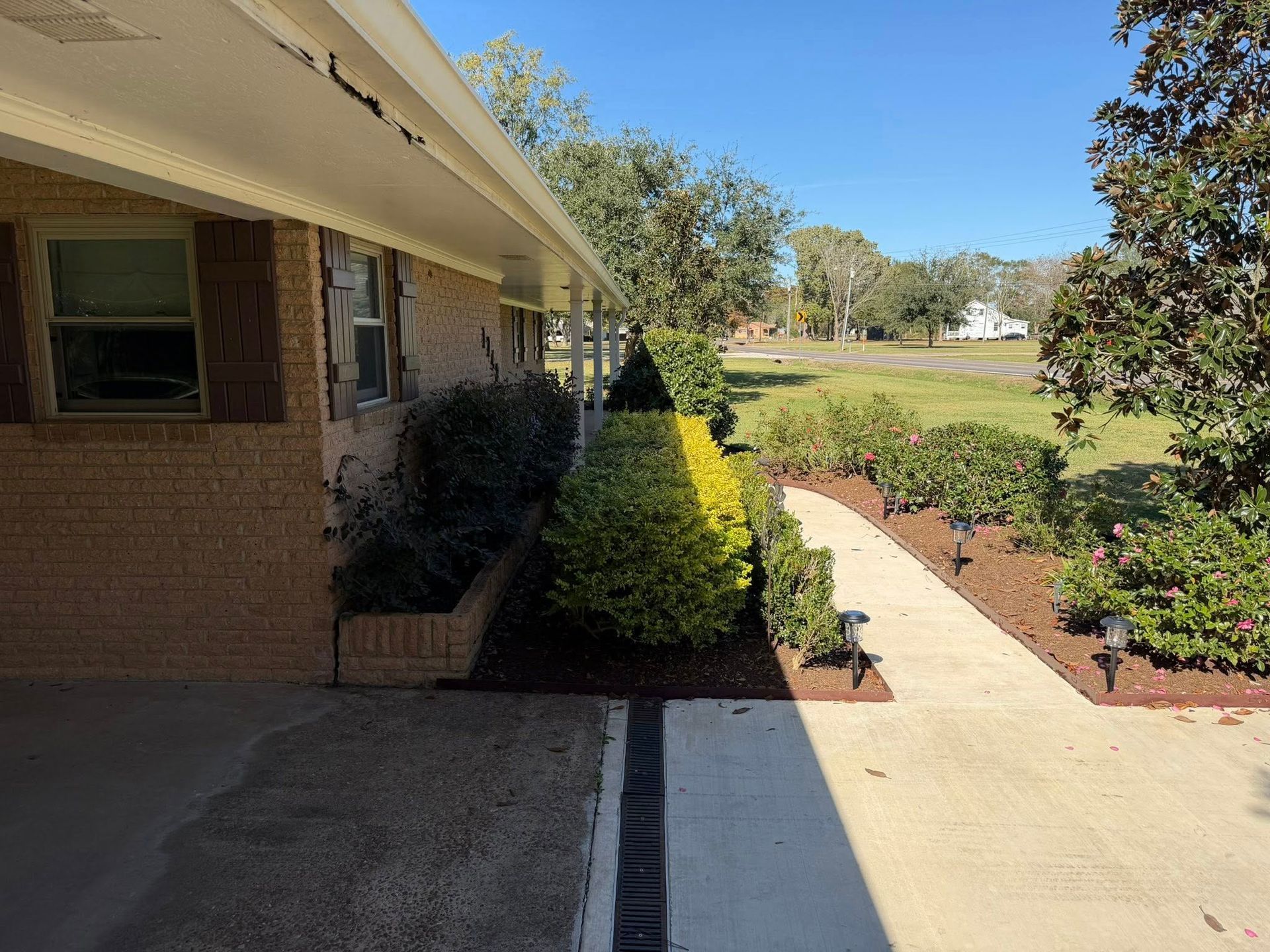Covered brick walkway beside a house, leading past shrubs to an open grassy yard on a sunny day