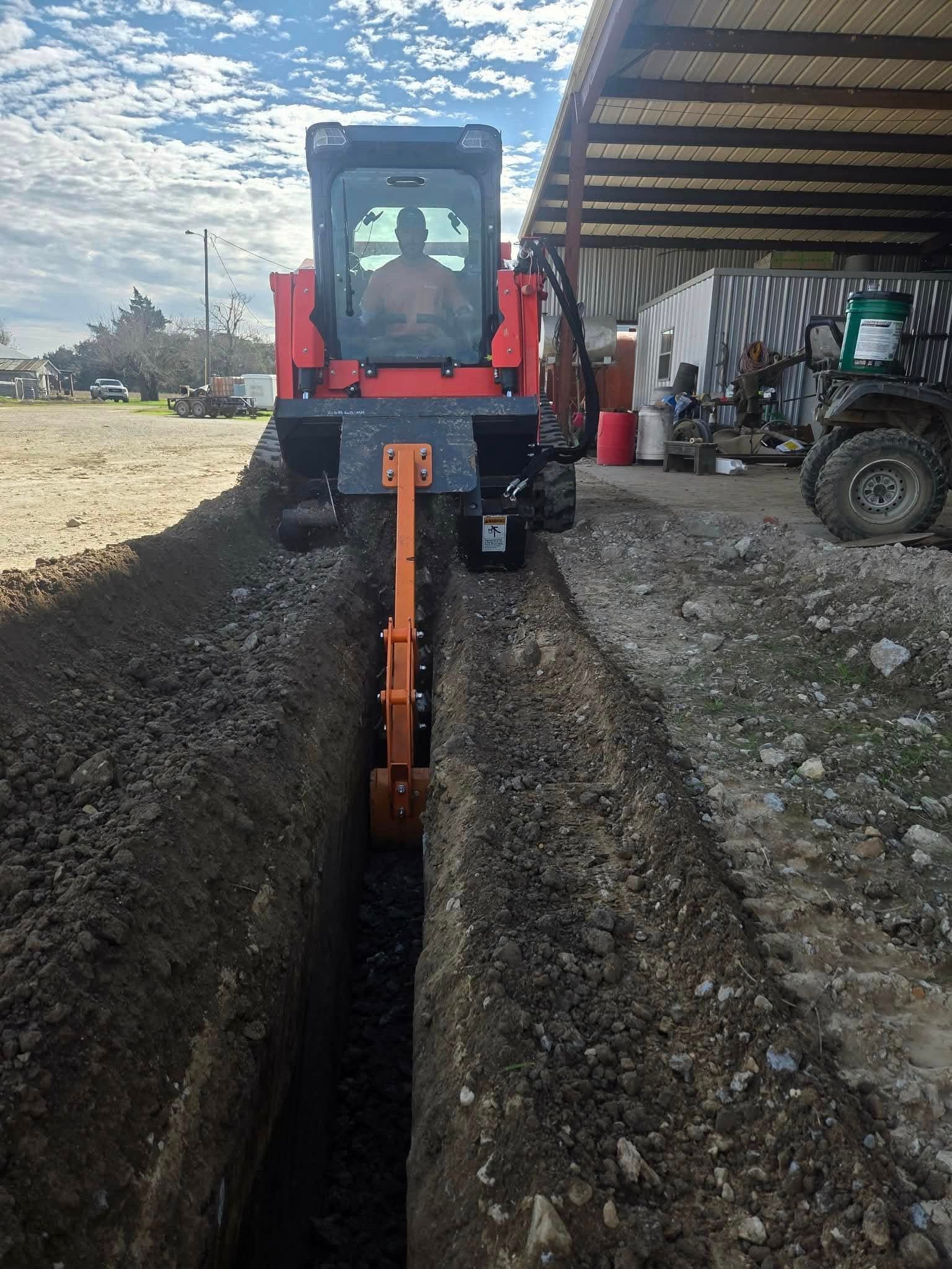 Red compact tractor digging a narrow trench beside a gravel path under a shed
