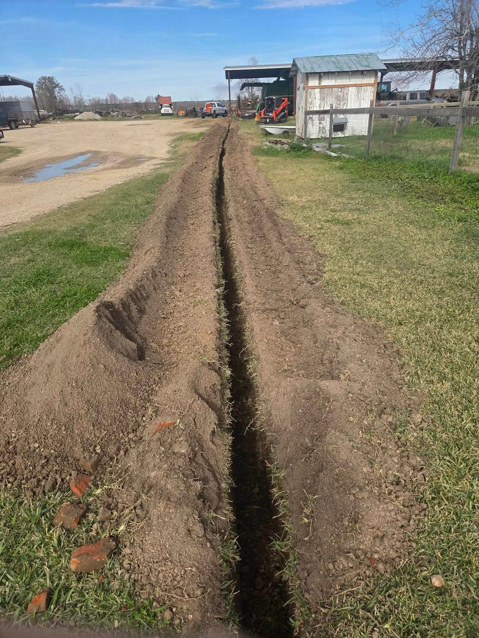Deep dirt trench running through a grassy field toward a small building and excavator in the distance