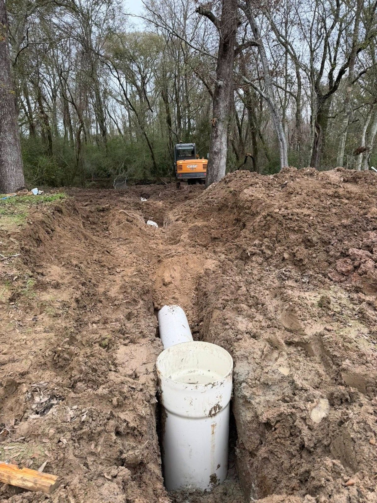 Excavator digging a muddy trench with a white pipe in the foreground and trees behind.