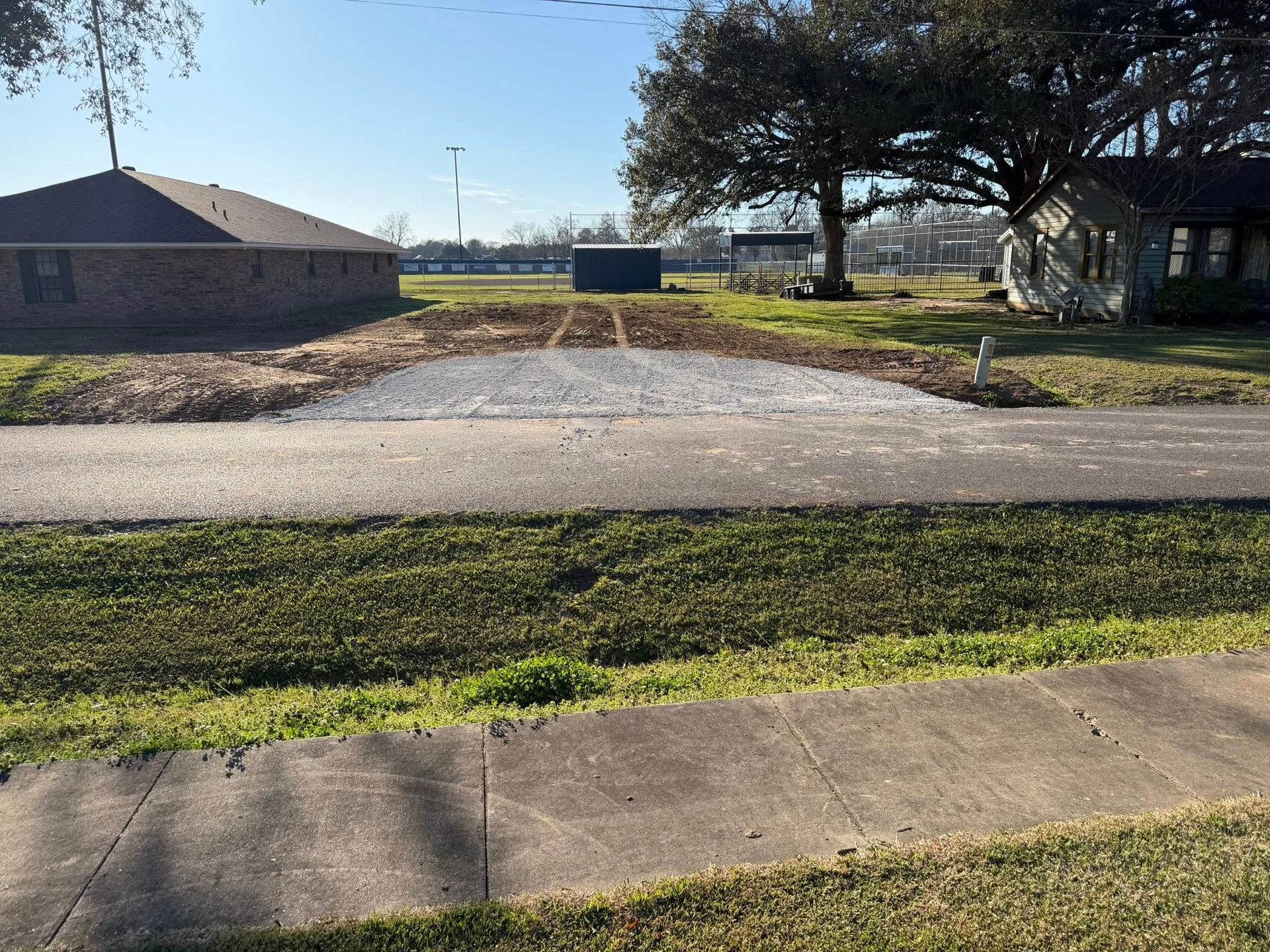 Empty gravel lot beside grassy curb and houses under a bright blue sky.