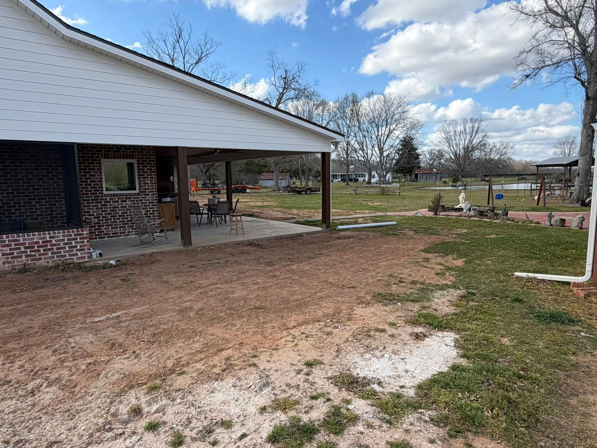Covered patio beside a muddy yard, with a brick house, picnic table, and trees under a cloudy sky