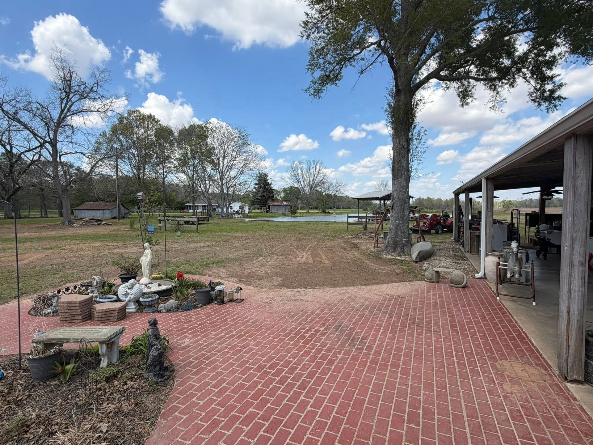 Brick patio beside a yard with trees, a small garden bed, and a covered porch under a blue sky.