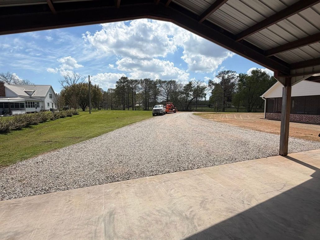 Gravel driveway under a metal carport with a red car in the distance, beside grassy field and houses.