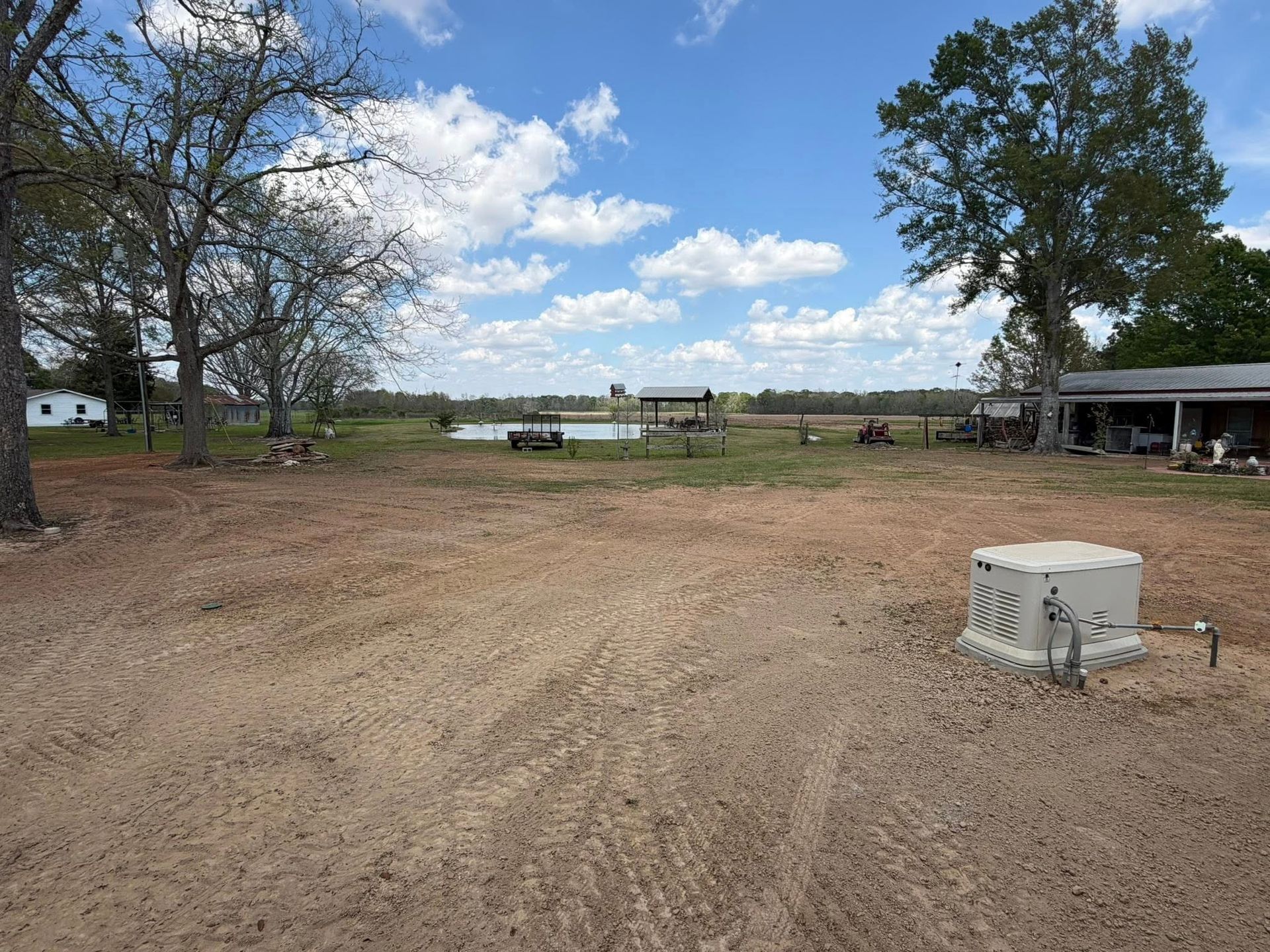 Dirt lot with a white cooler, trees, picnic table, and open field under a partly cloudy sky.
