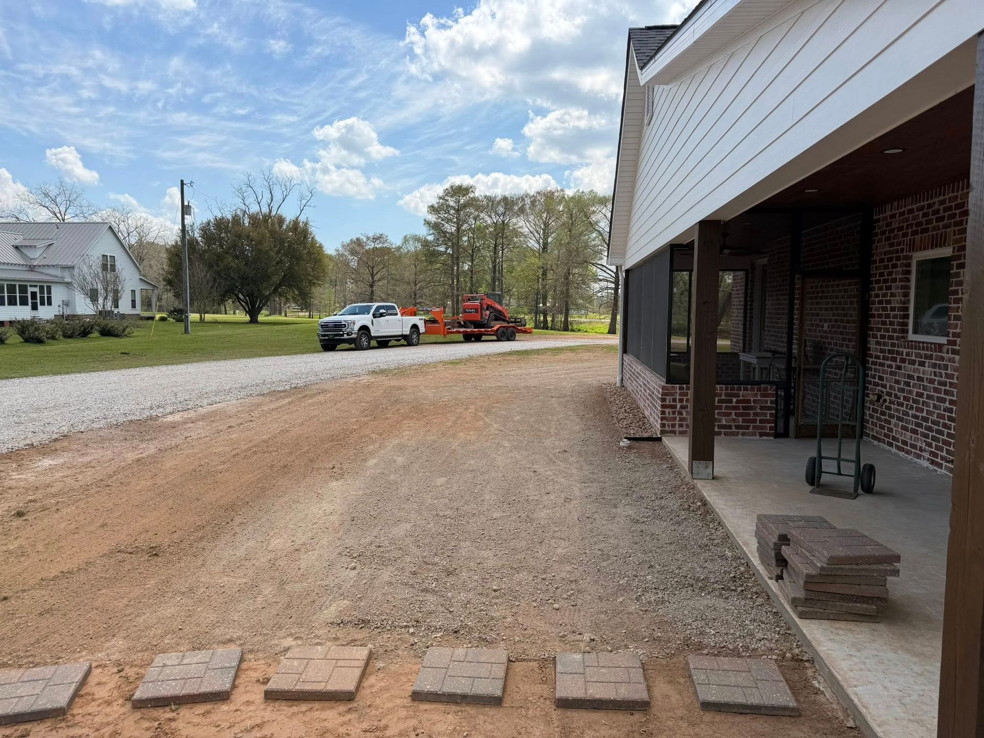 Gravel driveway beside a brick porch, with parked vehicles and a red tow truck in the distance under a blue sky
