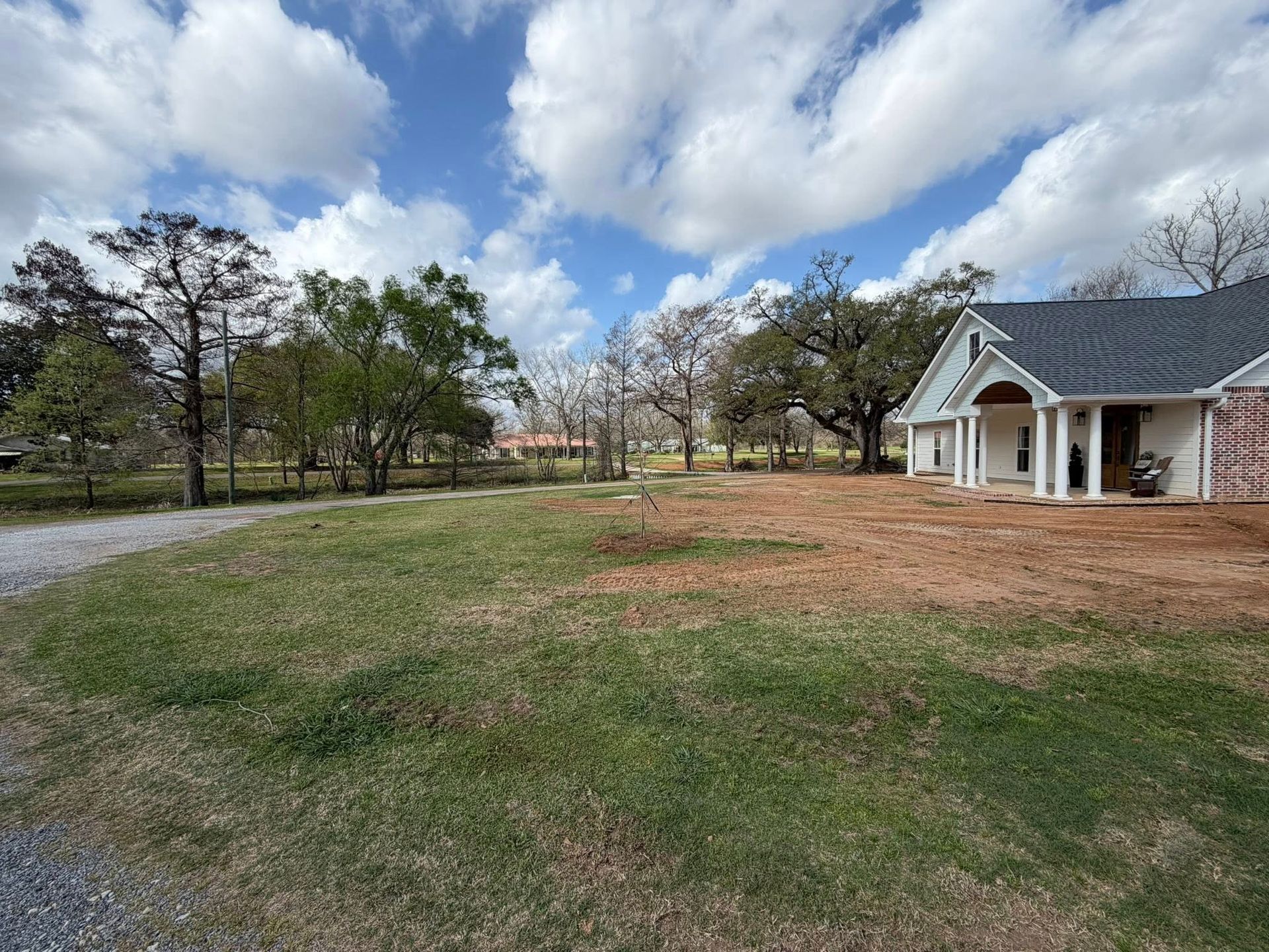 Suburban yard with grass, trees, and a white house under a cloudy blue sky