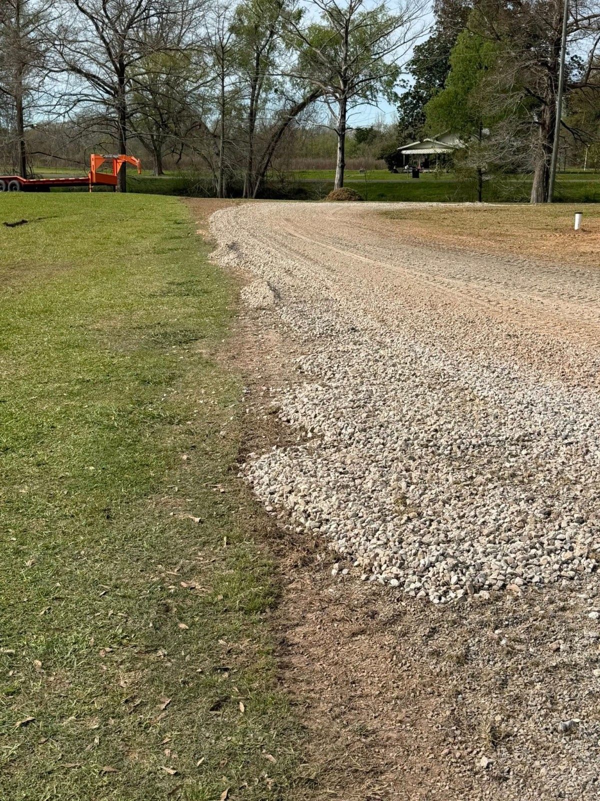 Gravel driveway curving through a grassy yard with trees and orange construction equipment in the background.