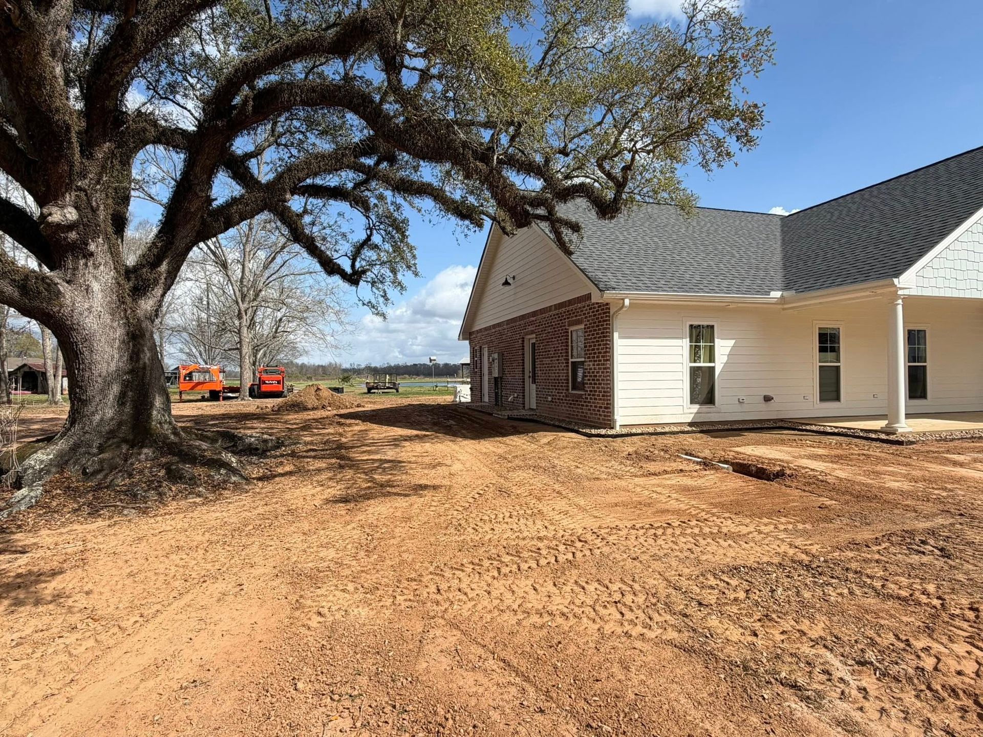 Rural farmhouse with large trees and bare dirt yard under a blue sky