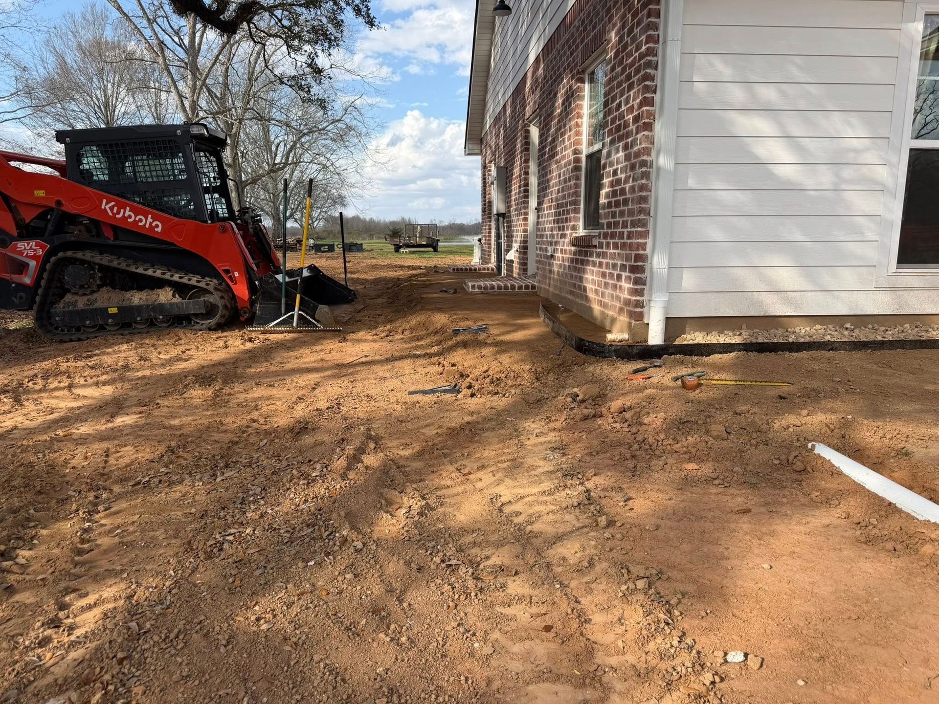 Red skid steer on dirt beside a brick-and-white house under construction.